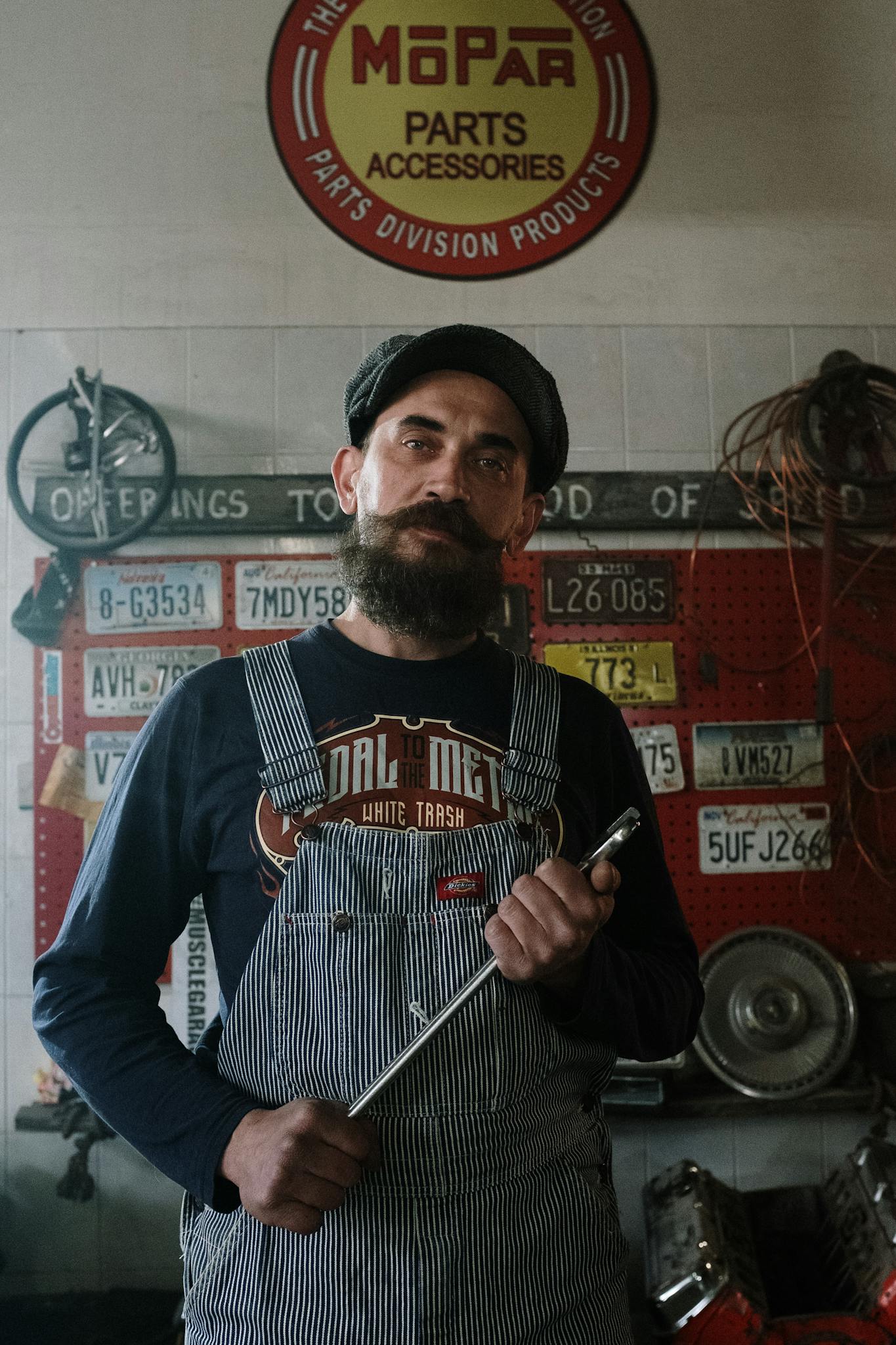 Bearded mechanic in overalls holding wrench in a garage adorned with license plates and vintage decor.